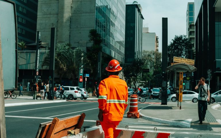 Trabalhador caminha por uma rua movimentada de São Paulo, cercado por pedestres e edifícios urbanos.